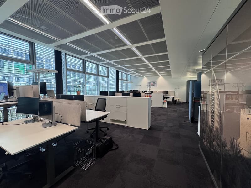 Modern office space with desks, chairs, multiple screens, laptops, and cabinets. Large windows provide natural light, and the room features a contemporary design with a mix of open and enclosed workspaces.