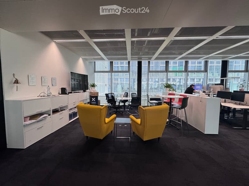 Modern office space with large windows, yellow chairs, a small table, desks, and computer setups.