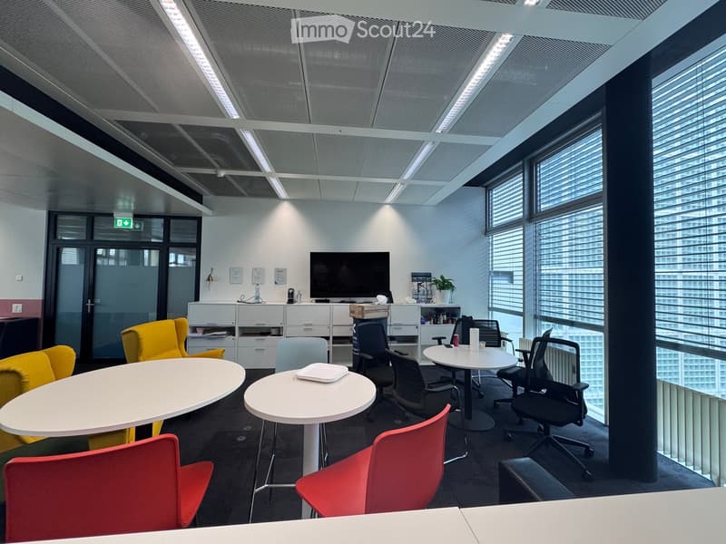 Modern office space with large windows, white tables, colorful chairs, and a TV screen.