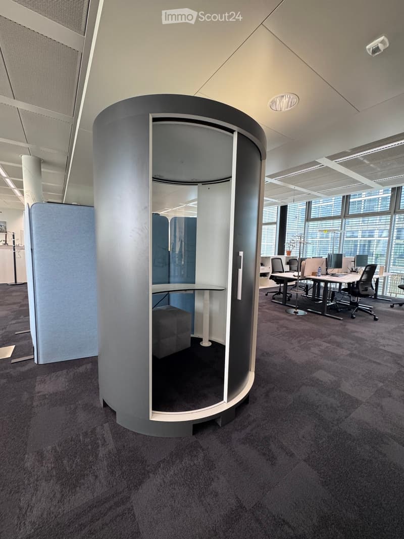 Modern office space with a round, enclosed workstation featuring a table, chair, and screen, surrounded by additional desks, chairs, and computer monitors in an open-plan area with large windows.