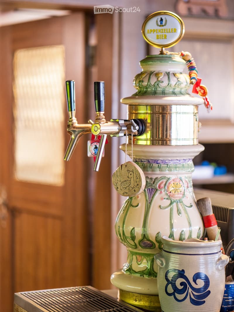 A beer tap with a decorative ceramic jug and mug, featuring a sign reading "Appenzeller Bier" and a small hanging ornament.