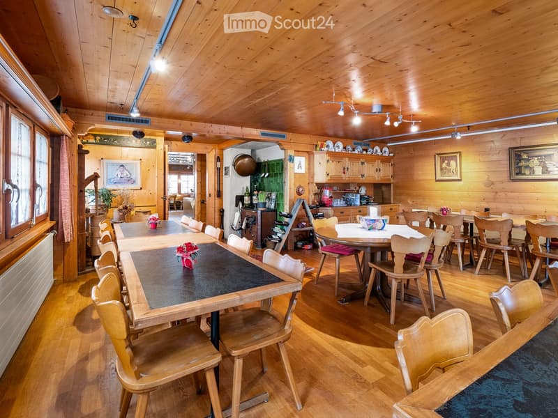 A spacious, wooden-paneled dining area with multiple tables and chairs, a kitchen area with cabinets and appliances, and decorative elements like framed pictures and flower vases.