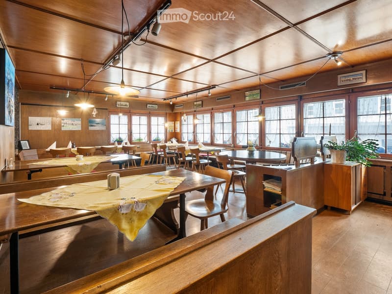 Restaurant interior with wooden tables, chairs, and large windows. Wooden ceiling and walls, pendant lights, and a potted plant on a counter.
