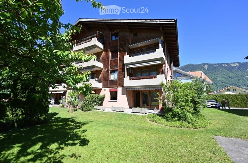 Multi-story wooden building with balconies, surrounded by a well-maintained lawn and trees, with a scenic mountain view in the background.