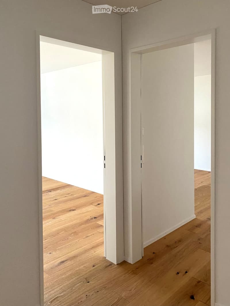 A modern, minimalist hallway with light wooden flooring and white walls, featuring an open doorway leading to another room.
