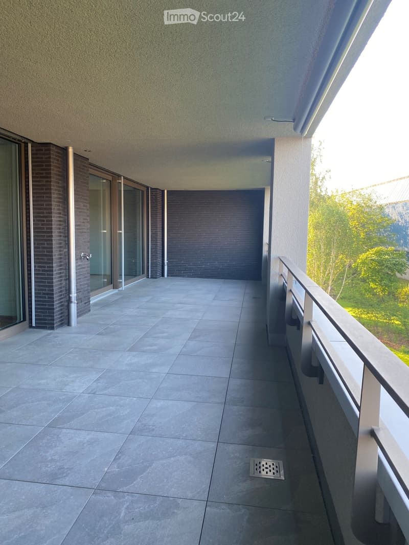 Modern balcony with tiled floor, glass sliding doors, metal railing, and a view of greenery.