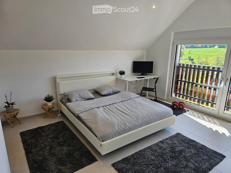 Modern bedroom with a white bed, grey bedding, a desk with a computer, a small stool, and a view of a green landscape through large windows.