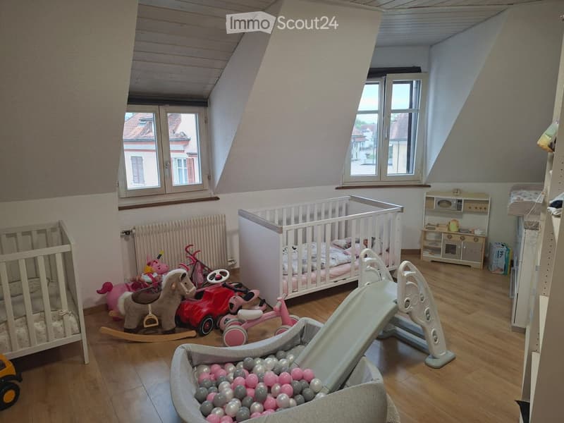 A well-lit attic bedroom with wooden flooring, two cribs, a variety of children's toys including a toy horse, toy car, toy bicycle, and a ball pit, along with a radiator and a small cabinet.