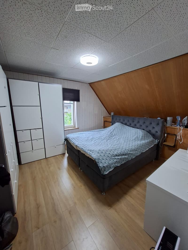 Cozy attic bedroom with wooden flooring, slanted wooden ceiling, large window, grey upholstered bed, white wardrobe, and nightstand with lamp.