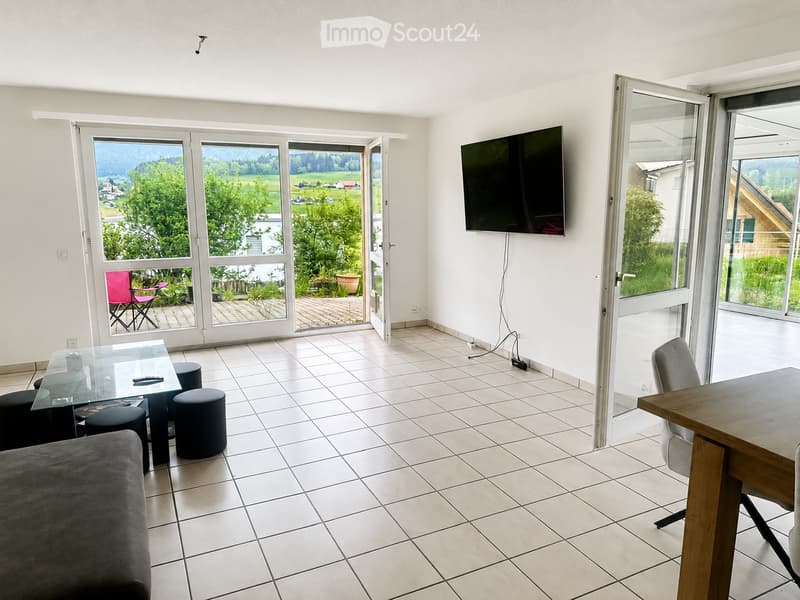 Bright living room with tiled floor, large windows and sliding doors leading to a balcony, a sofa, a glass table with stools, a chair, and a television mounted on the wall.