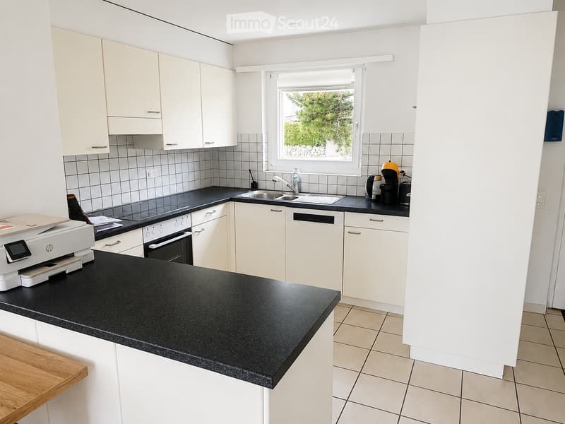 Modern kitchen with white cabinets, black countertops, and stainless steel appliances including a dishwasher, oven, and coffee maker. A printer sits on the counter, and a window offers a view of greenery outside.