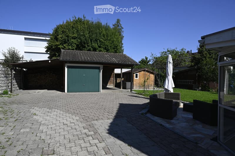 Garden area with a paved brick pathway, outdoor seating area with a sofa and umbrella, garage, and surrounding greenery.