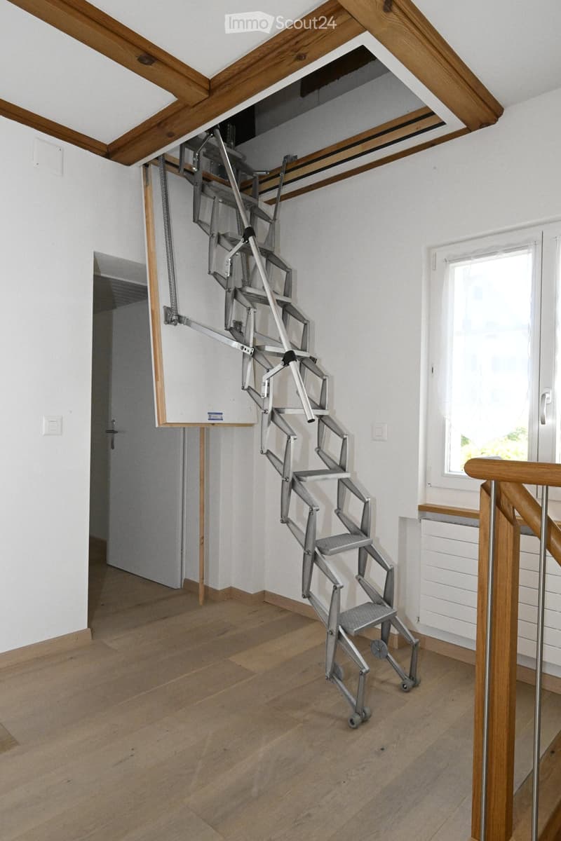 An unfurnished room with wooden flooring, white walls, and a retractable metal staircase leading to an attic space. The room features a window and a door leading to another area.
