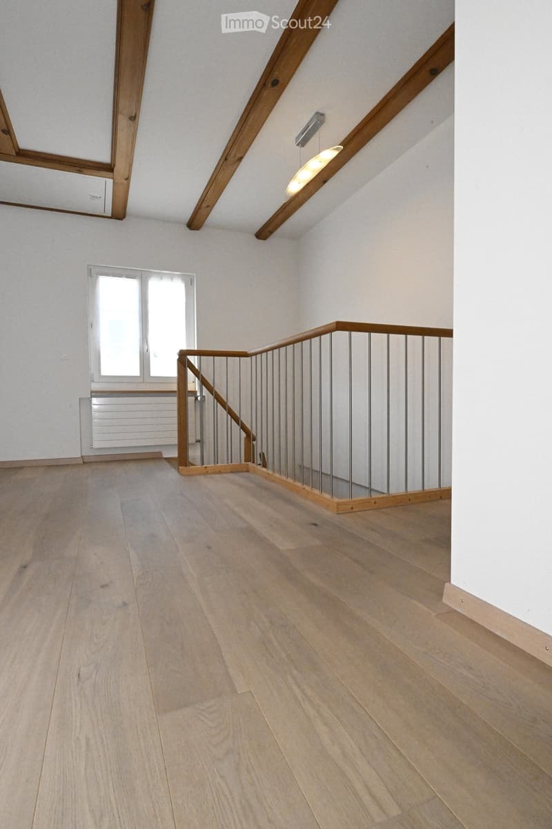 Empty hallway with wooden flooring, white walls, and a staircase with wooden railings and metal balusters. A window and radiator are visible on the left side.