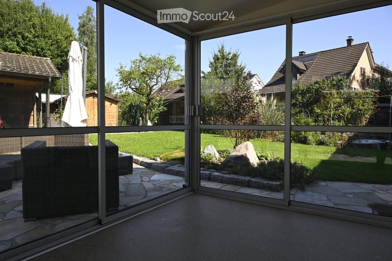 A spacious, well-maintained garden visible through large glass doors, featuring a stone pathway, a large rock, neatly trimmed grass, and surrounding trees and shrubs.