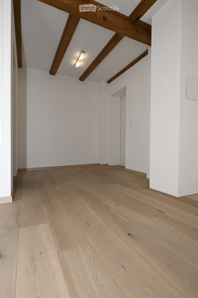 Empty room with light wooden flooring and white walls, featuring exposed wooden beams on the ceiling and a small recessed light fixture.