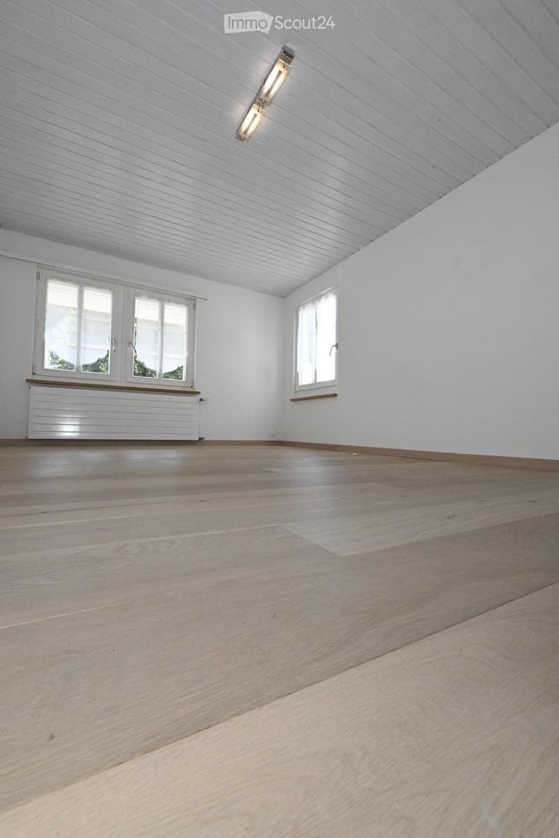 Empty room with light wooden flooring, white walls, and white ceiling with wooden paneling. Two windows with white frames and blinds, and a white radiator under one window.