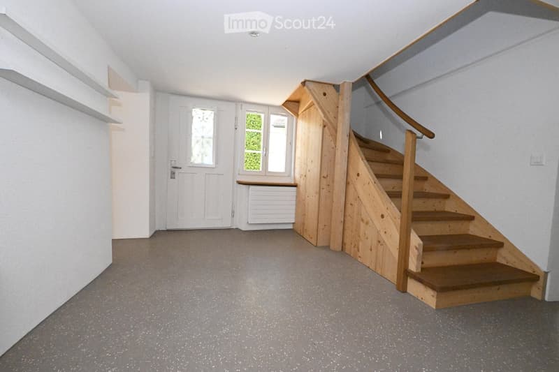Empty hallway with a wooden staircase leading upwards, white walls, grey carpeted floor, and a door with glass panels.