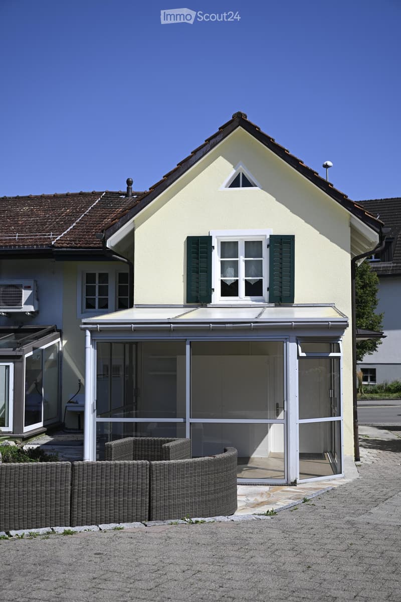 Two-story house with beige exterior, green shutters, and a covered patio area with wicker furniture.