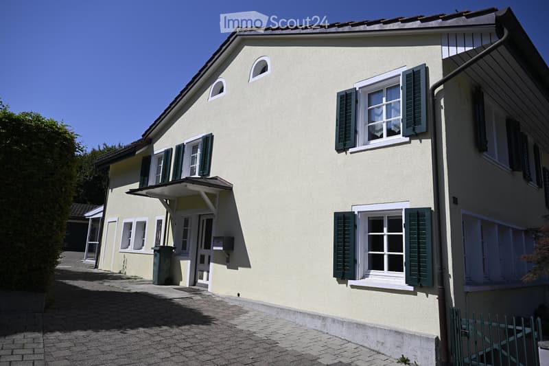 Two-story house with light yellow walls, white-framed windows with green shutters, and a small covered entrance area.