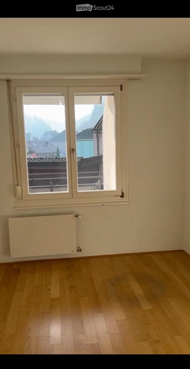 Empty room with wooden flooring, large window with double doors leading to a balcony, radiator below the window, and a view of rooftops and mountains.