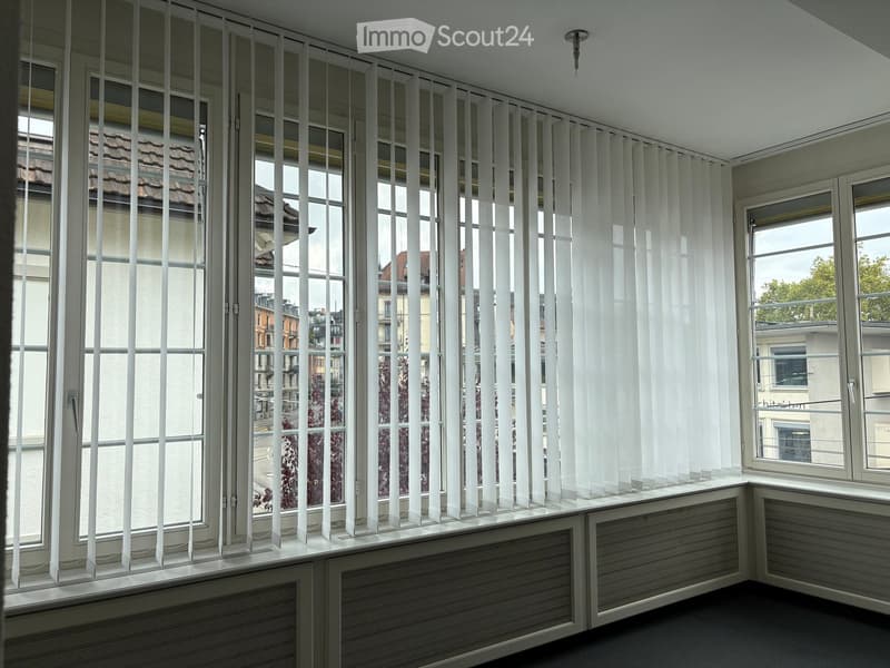 Empty room with large windows covered by vertical blinds, radiator under the windows, and a view of the cityscape.