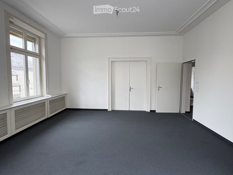 Empty room with white walls, grey carpet, large window with radiator cover, and two doors.