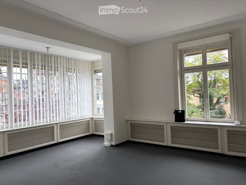 Empty room with grey carpeted floor, white walls, large windows with blinds, radiators, and a small black bag on the windowsill.