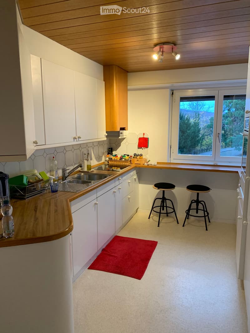 Modern kitchen with white cabinets, wooden countertops, a sink, and two stools. Features a red rug on the floor and a window with a view of greenery.
