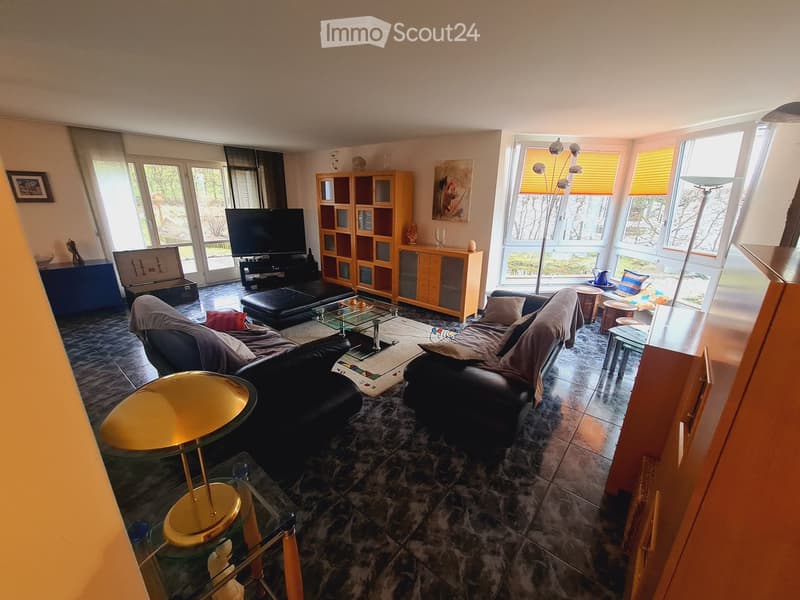 Modern living room with large windows, dark tiled floor, two black leather sofas, glass coffee table, TV, wooden cabinet, and decorative items.