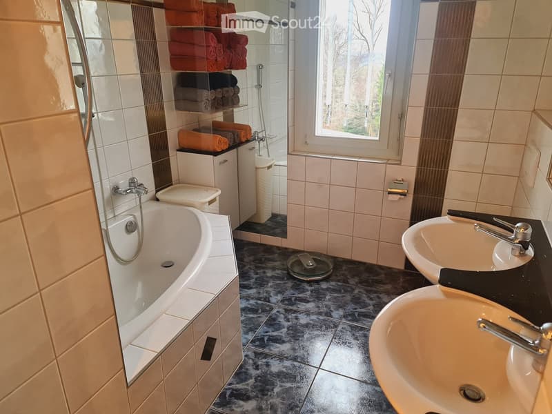 Modern bathroom with a bathtub, double sink, and a large mirror. Features tiled walls, a window, and neatly stacked towels on a shelf.