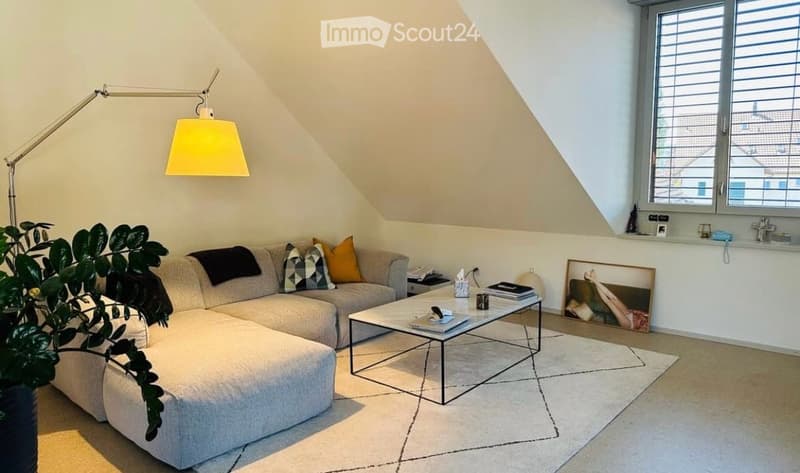 Modern living room with a grey sectional sofa, coffee table, floor lamp, and a large window with blinds. The room features a slanted ceiling and a small plant in the corner.
