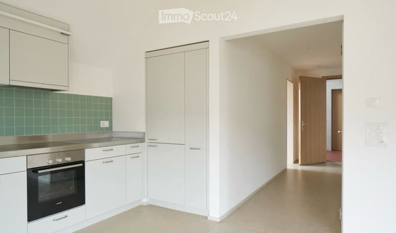 Modern kitchen with white cabinets, stainless steel oven, and green tiled backsplash. Open doorway leads to another room.