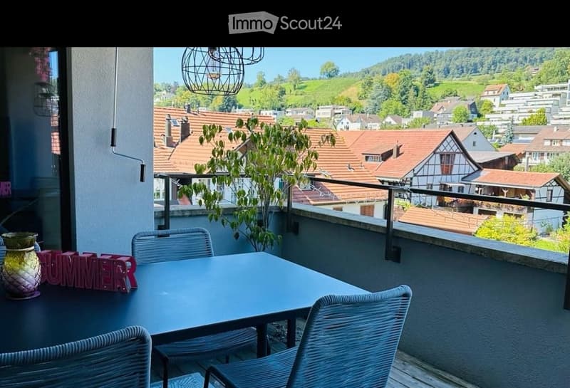 Balcony with a scenic view of a town and hills, featuring a table, chairs, and a decorative vase.