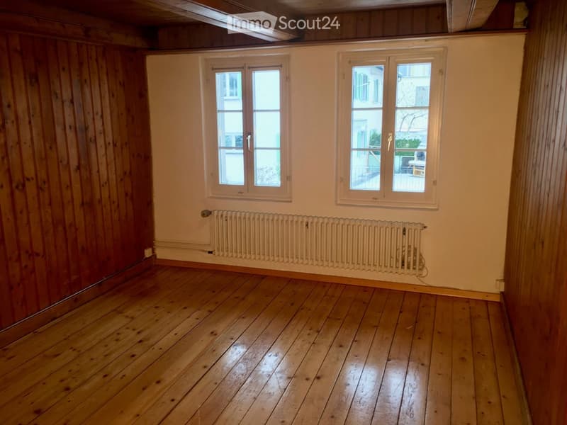 Empty room with wooden flooring, white walls, and two windows with white frames. A radiator is mounted on the wall below the windows.