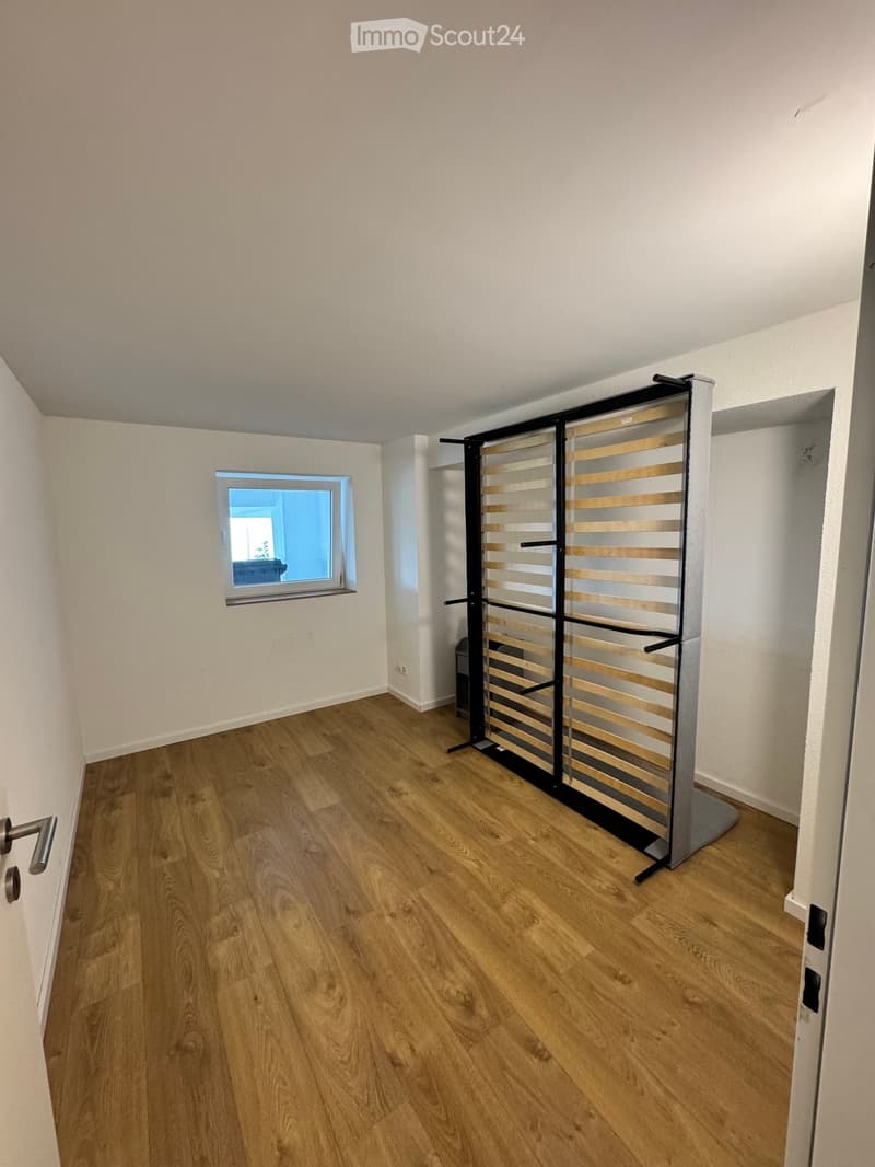 Empty room with wooden flooring, white walls, and a window with a view of another room. A clothes drying rack is positioned near the window.