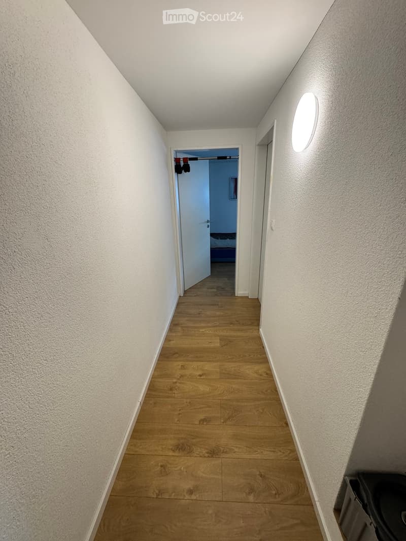 A narrow hallway with light wooden flooring and white walls, featuring a round wall-mounted light fixture and an open door leading to another room.