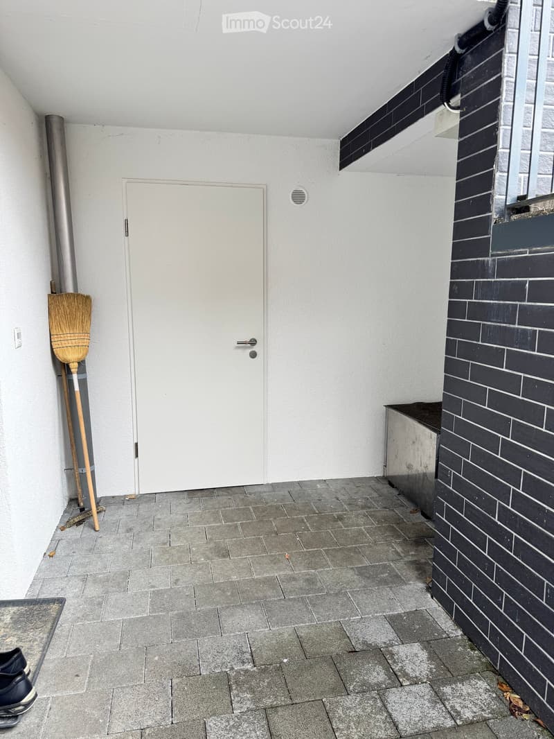 A narrow hallway with a tiled floor, white walls, and a broom leaning against the wall. A white door is visible at the end of the hallway.
