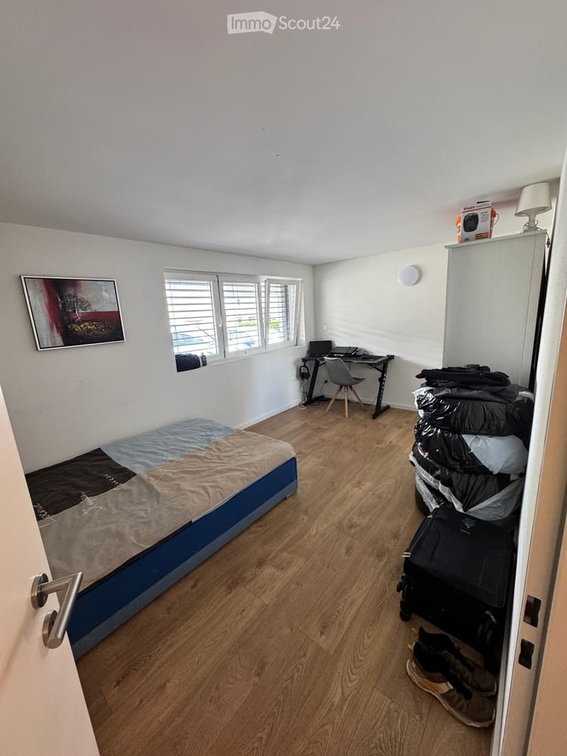 A small, furnished bedroom with a single bed, desk, chair, and a suitcase. The room features wooden flooring, white walls, and a window with blinds.