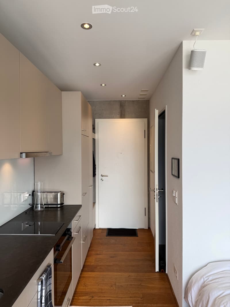 Modern kitchen with light wood flooring, white cabinets, black countertops, and a small hallway leading to a bedroom with a white bed.