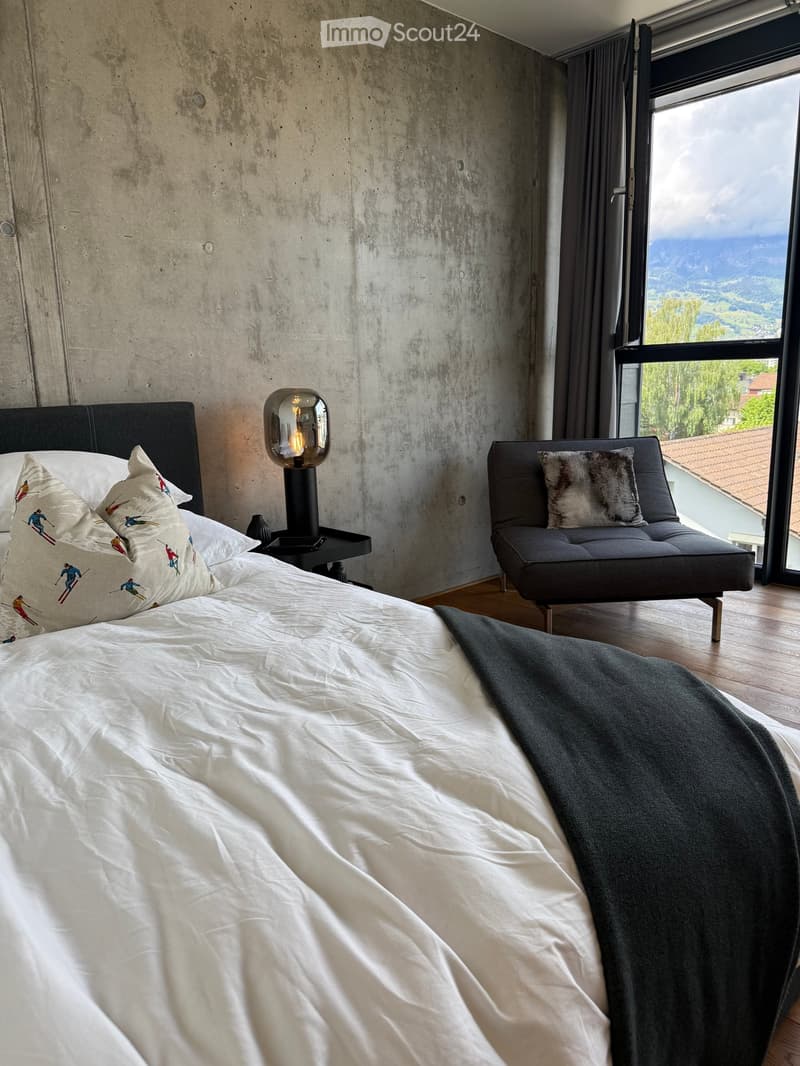Modern bedroom with a large bed, white bedding, decorative pillows, a sleek black chair, and a contemporary table lamp. The room features exposed concrete walls and large windows with a scenic mountain view.