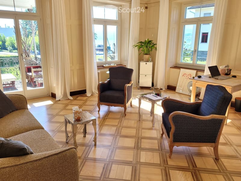 Bright living room with large windows and a view, featuring a sofa, two armchairs, a coffee table, a desk with a laptop, and a small plant on a side table.