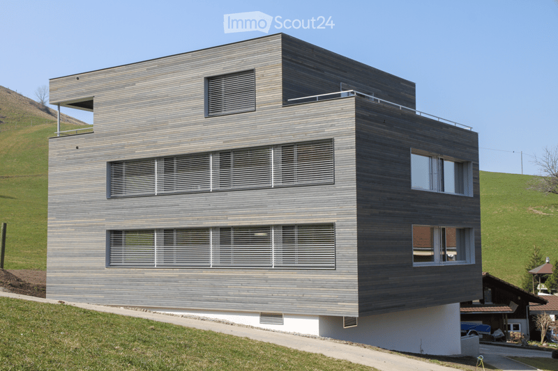 Modern architecture, wooden facade, multiple windows, balcony on top floor, white lower section, on a hill.