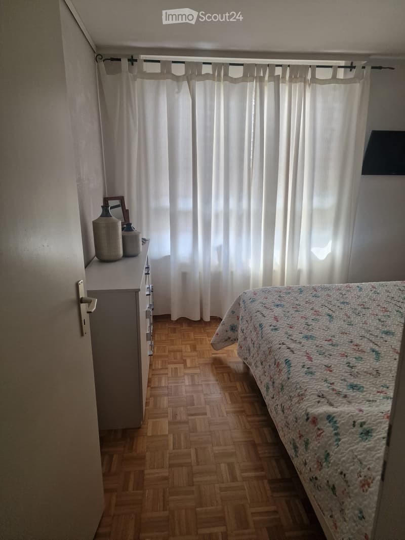 Bedroom with wooden herringbone flooring, a bed with a floral quilt, a nightstand with vases and a mirror, and white curtains covering a window.