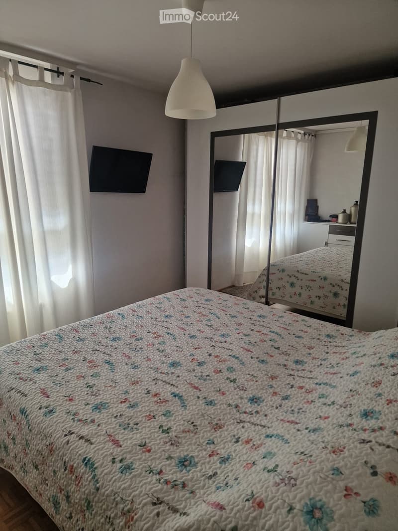 A well-lit bedroom with a large bed covered in a floral quilt, a wall-mounted TV, a large mirror reflecting part of the room, and a hanging lamp above the bed.
