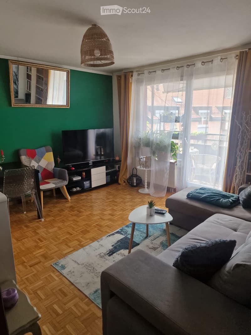 Modern living room with wooden flooring, large windows, a grey sectional sofa, a colorful armchair, a coffee table, a TV stand, and a hanging woven lamp.
