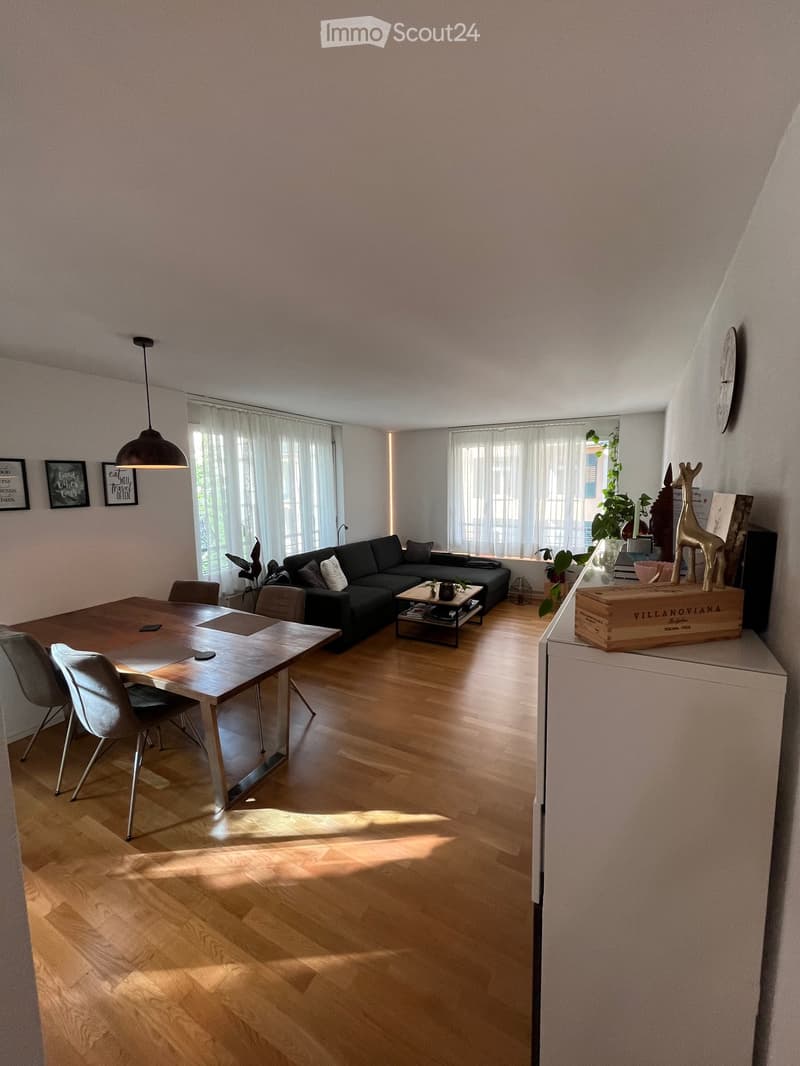 Bright living and dining area with wooden flooring, large windows with white curtains, a black sectional sofa, a wooden coffee table, a dining table with chairs, and a pendant light fixture.