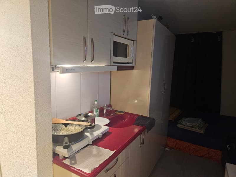 Compact kitchen with white cabinets, red countertop, microwave, stove, sink, and various kitchen items.