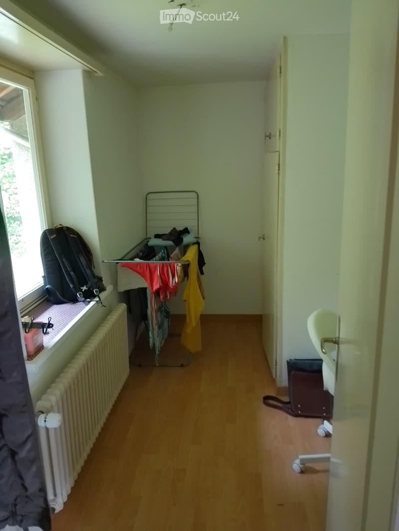 A narrow hallway with wooden flooring, a radiator along one wall, a clothes rack with hanging garments, a backpack on a windowsill, and a suitcase near the door.