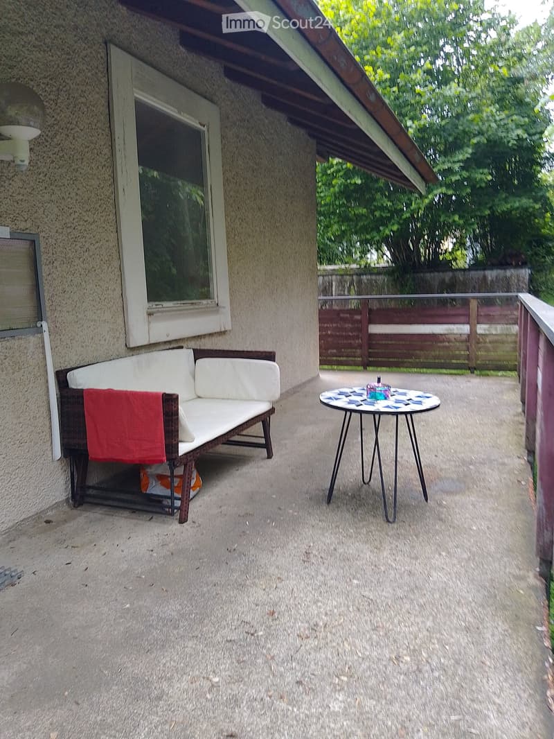 Balcony with a white sofa, red cushion, small round table with a patterned top, and a glass railing overlooking a fenced garden.